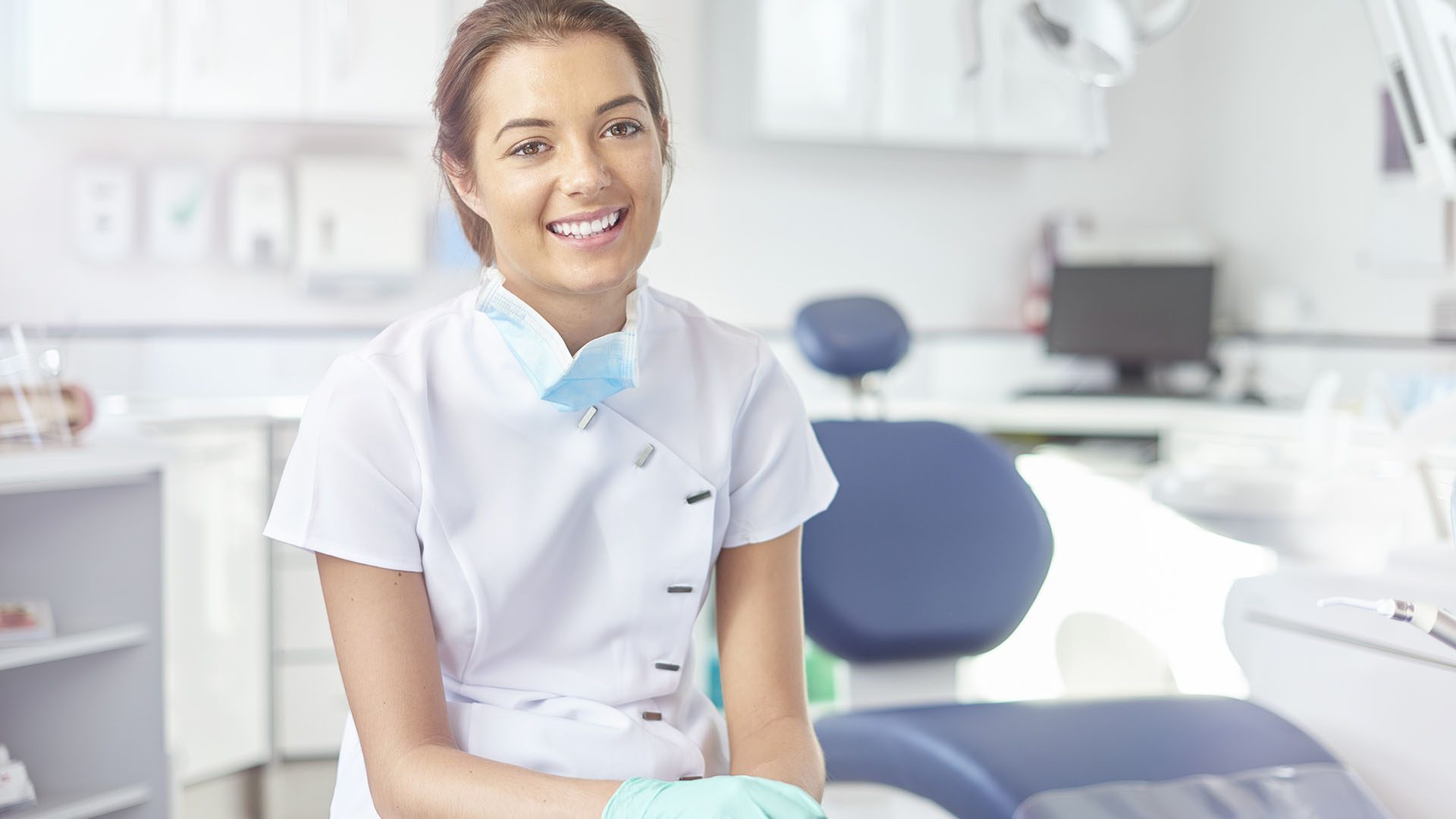 A young female dental nurse is looking proudly to camera from her dentist's office. In the background a modern dentist surgery can be seen brightly lit .